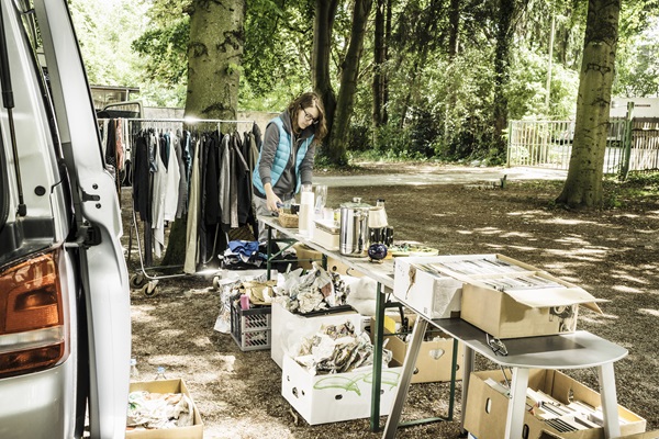 Rommelmarkt in een bosrijke omgeving, met een kraam vol tweedehands spullen, boeken en kleding. Een verkoper staat achter de tafel en schikt de items.