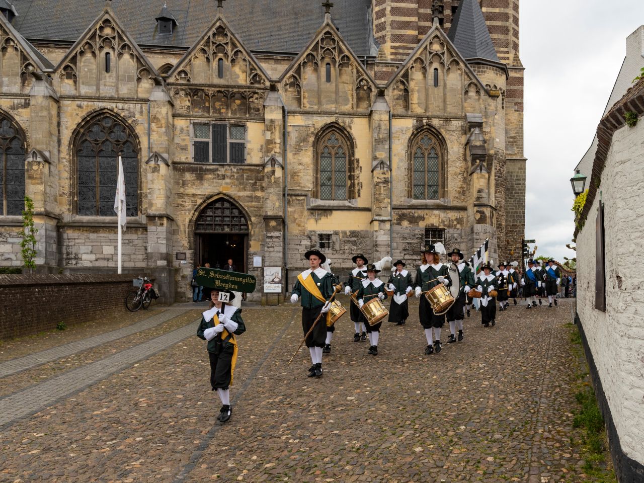Optocht van een schuttersgilde in traditionele kledij door de historische straten van Thorn, met op de achtergrond de imposante Abdijkerk.