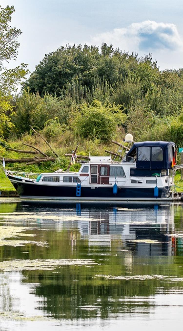 Een motorboot ligt aangemeerd langs een groene oever in een rustige, waterrijke omgeving. Een vrouw zit op een stoel aan de waterkant. Met de boot naar Limburg geniet je van rust, natuur en groene oevers.