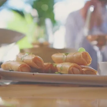 Bord met versgebakken loempia’s op een tafel in een restaurant, met een glas wijn en een persoon op de achtergrond.