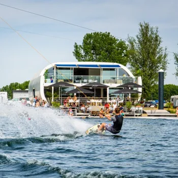 Wakeboarder op een cable park voor een modern paviljoen met terras aan het water, omringd door toeschouwers.
