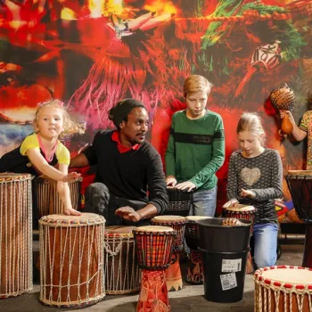 Man begeleidt drie kinderen tijdens een interactieve djembéworkshop met kleurrijke achtergrond.