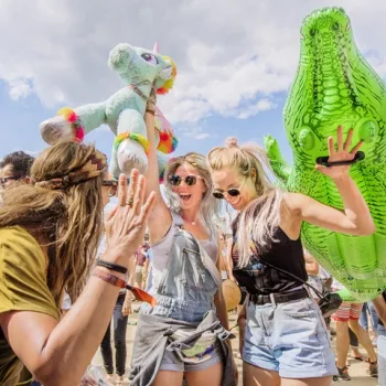 Twee vrouwen feesten met grote opblaasfiguren tussen het festivalpubliek onder een blauwe lucht.